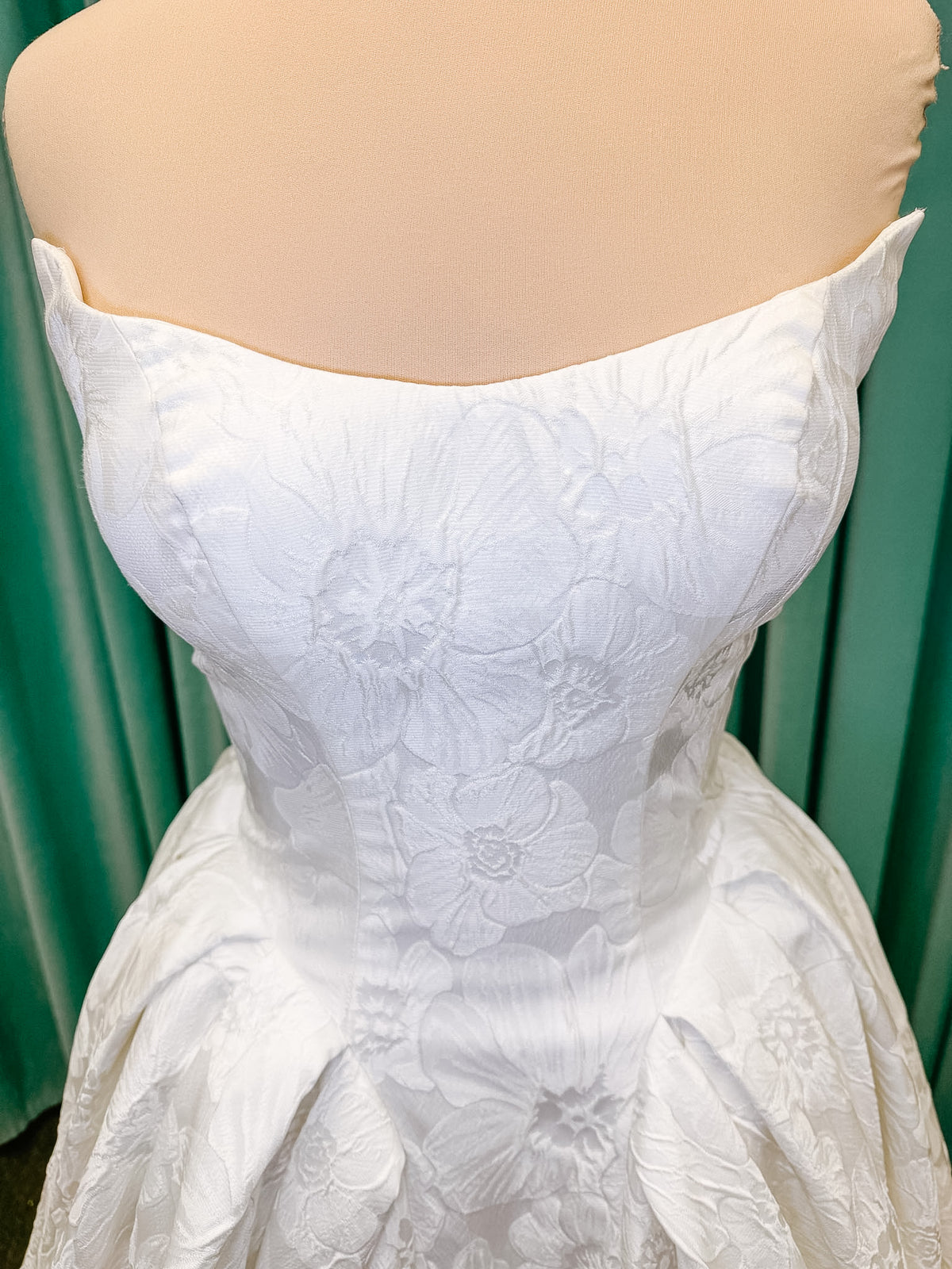 Haley Mai Bridal Lorea ballgown wedding dress on mannequin with strapless scoop neckline, seamed basque waist bodice, pleated ivory floral jacquard skirt, and long train