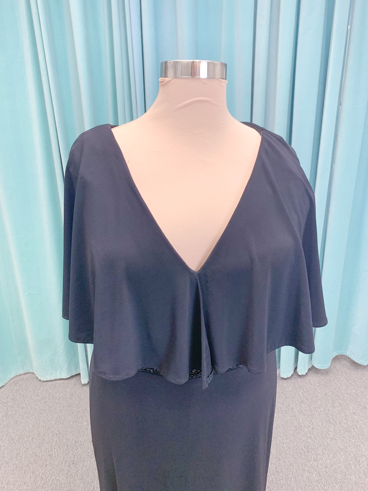Morilee MLNY 73124 sheath mother of the bride dress on mannequin with V-neckline, flowing capelet overlay, crystal beaded waistband, and slim jersey sheath skirt