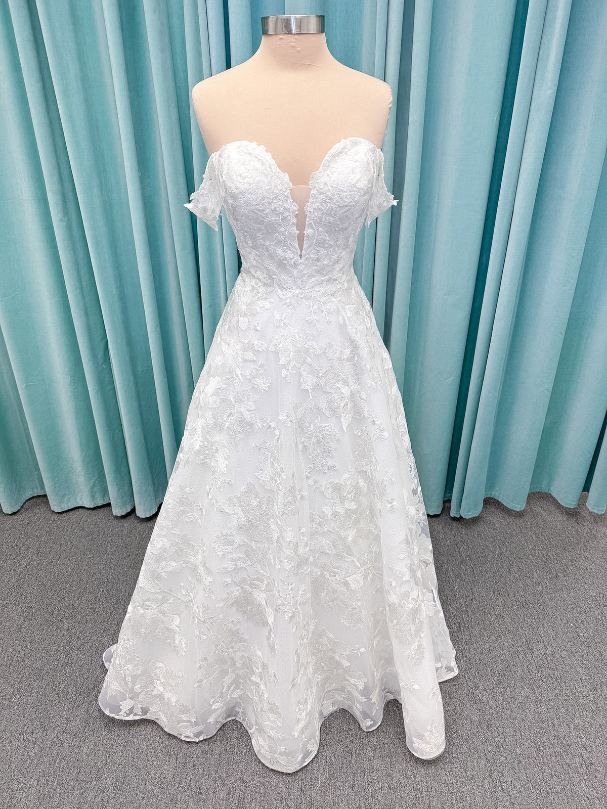 Moonlight Bridal J6818 Channary A-line wedding dress on mannequin with sweetheart illusion neckline, large floral embroidered lace with sequins, off-the-shoulder lace swag sleeves, full net skirt, and chapel train