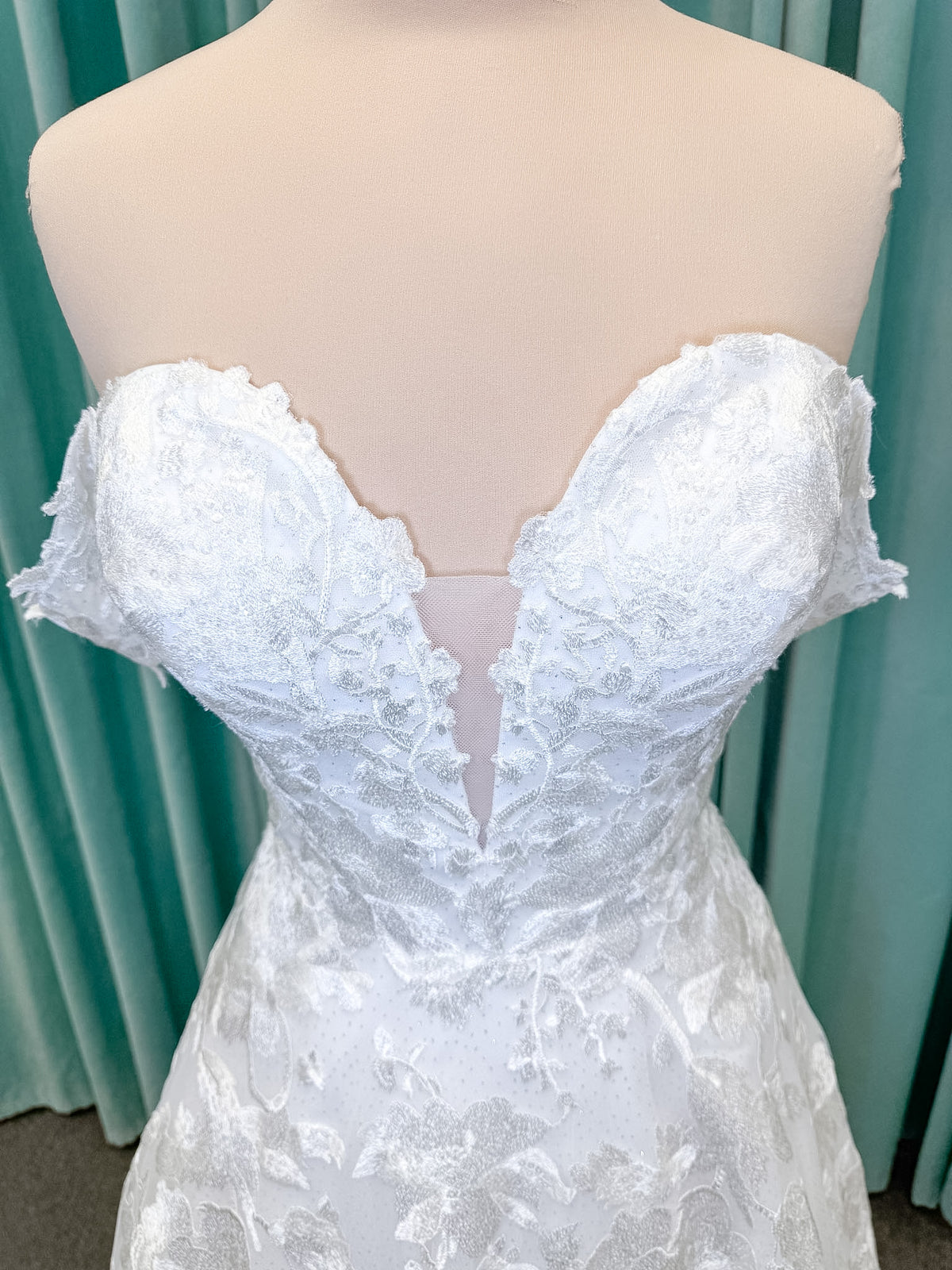 Moonlight Bridal J6818 Channary A-line wedding dress on mannequin with sweetheart illusion neckline, large floral embroidered lace with sequins, off-the-shoulder lace swag sleeves, full net skirt, and chapel train