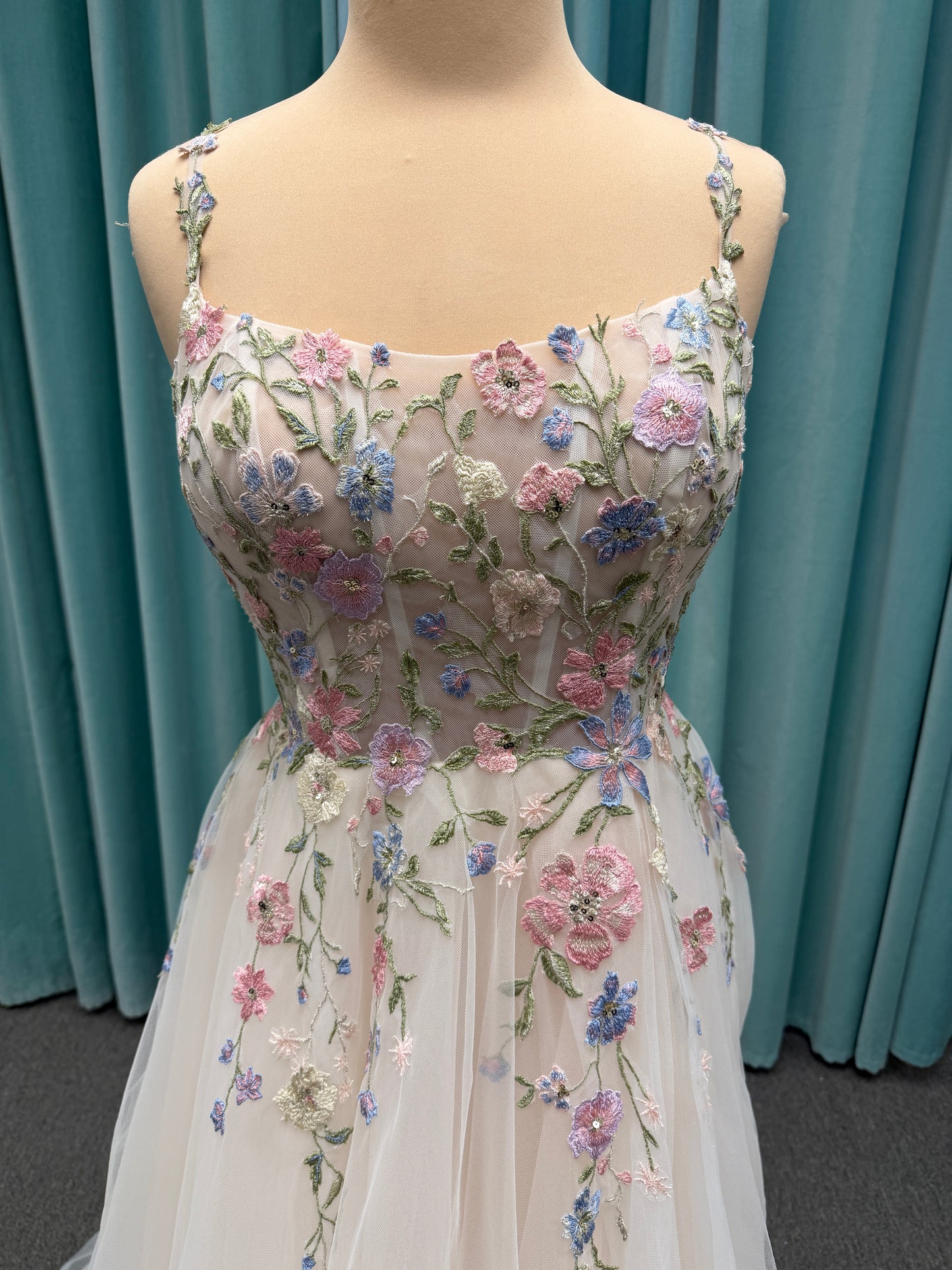 Stella York 8063 ivory and pastel colorful embroidered lace A-line wedding dress on mannequin with floral spaghetti straps, scoop neckline, and soft tulle skirt.
