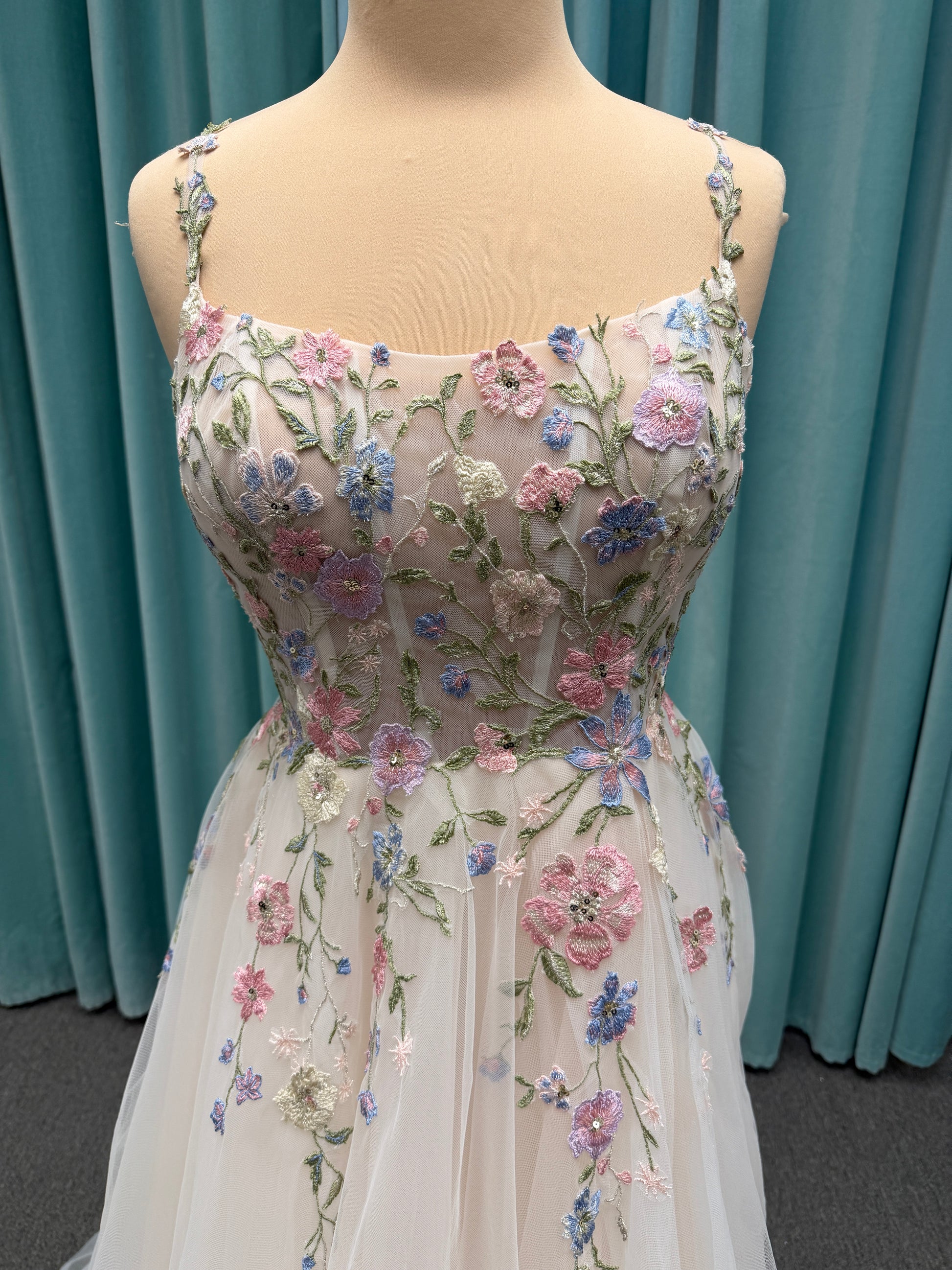 Stella York 8063 ivory and pastel colorful embroidered lace A-line wedding dress on mannequin with floral spaghetti straps, scoop neckline, and soft tulle skirt.
