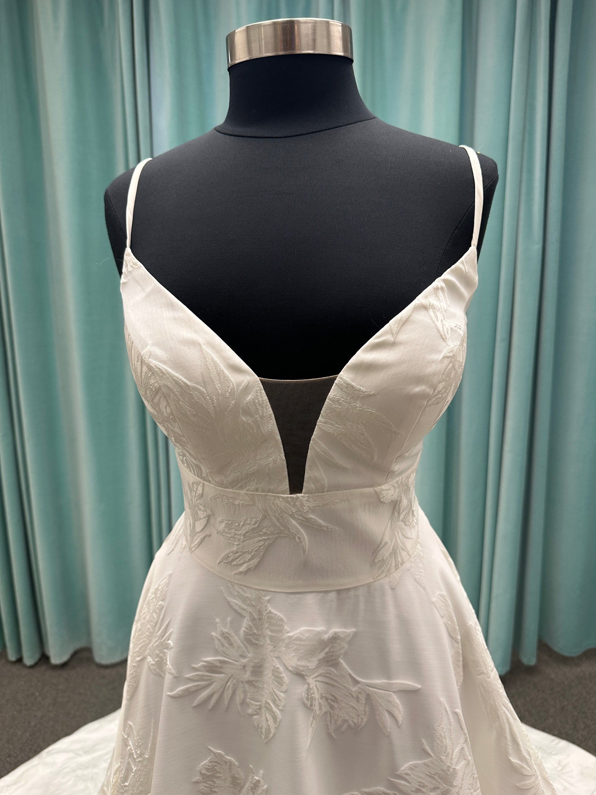 Lisha ivory spaghetti strap V-neck ballgown wedding dress on mannequin in floral jacquard fabric with a full sweeping skirt.
