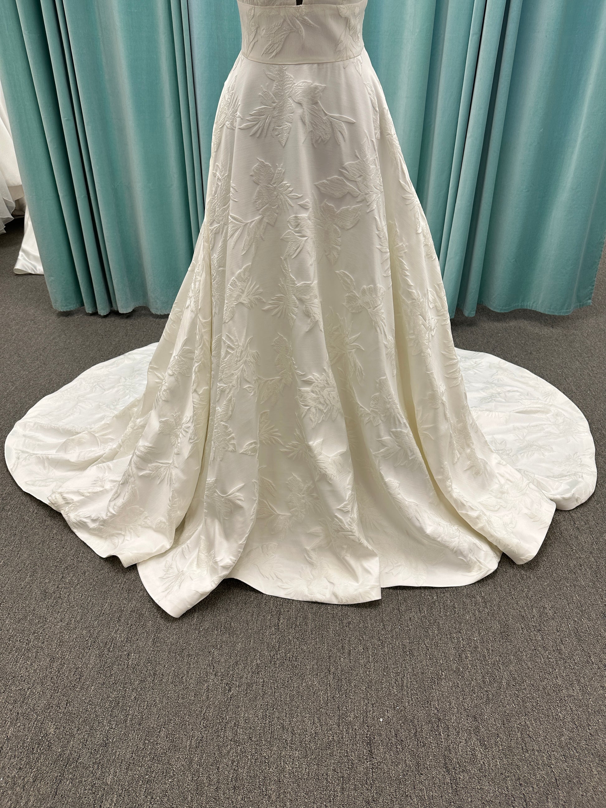Lisha ivory spaghetti strap V-neck ballgown wedding dress on mannequin in floral jacquard fabric with a full sweeping skirt.
