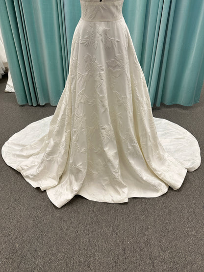 Lisha ivory spaghetti strap V-neck ballgown wedding dress on mannequin in floral jacquard fabric with a full sweeping skirt.
