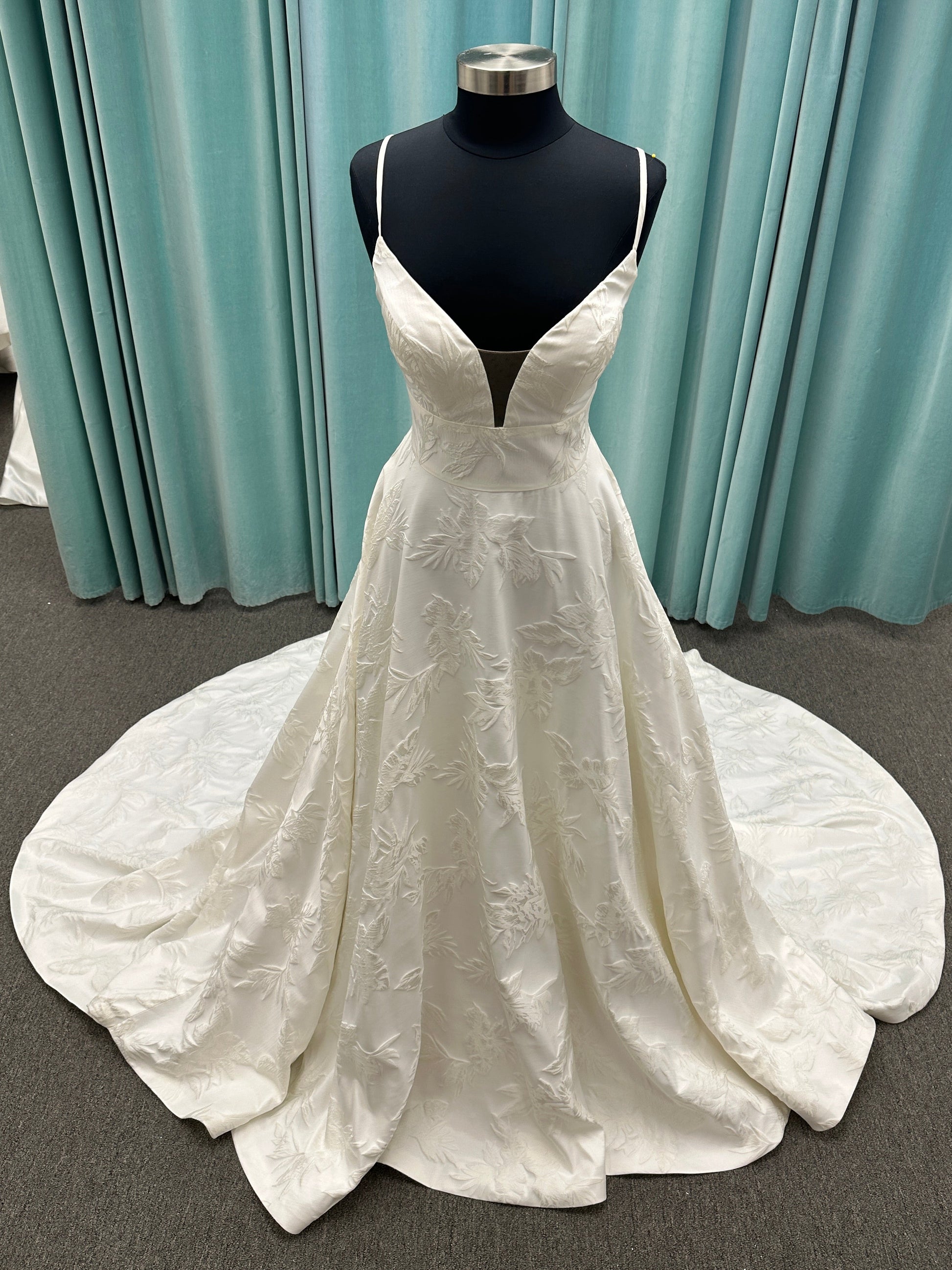 Lisha ivory spaghetti strap V-neck ballgown wedding dress on mannequin in floral jacquard fabric with a full sweeping skirt.
