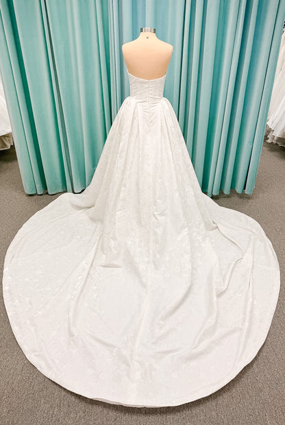 Haley Mai Bridal Lorea ballgown wedding dress on mannequin with strapless scoop neckline, seamed basque waist bodice, pleated ivory floral jacquard skirt, and long train
