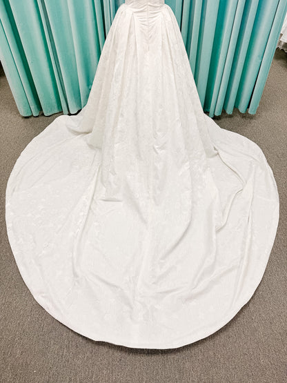 Haley Mai Bridal Lorea ballgown wedding dress on mannequin with strapless scoop neckline, seamed basque waist bodice, pleated ivory floral jacquard skirt, and long train