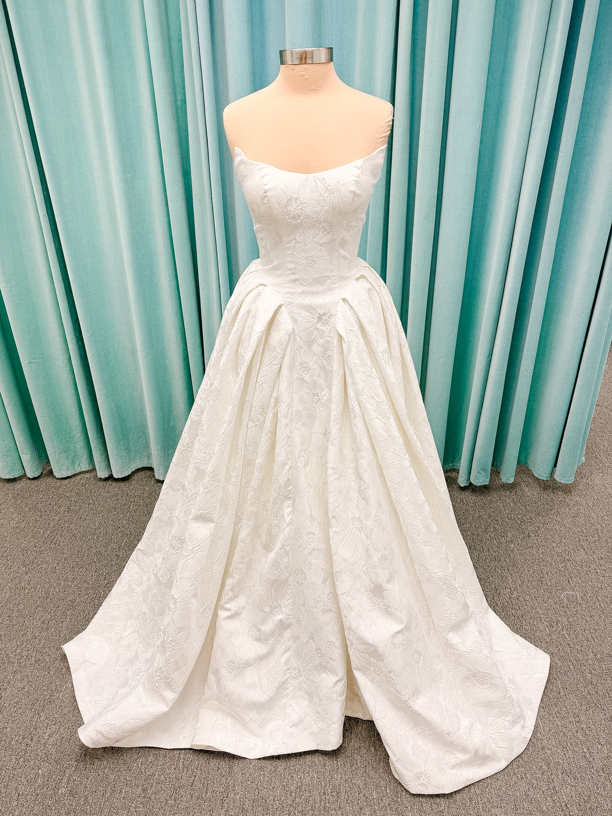 Haley Mai Bridal Lorea ballgown wedding dress on mannequin with strapless scoop neckline, seamed basque waist bodice, pleated ivory floral jacquard skirt, and long train