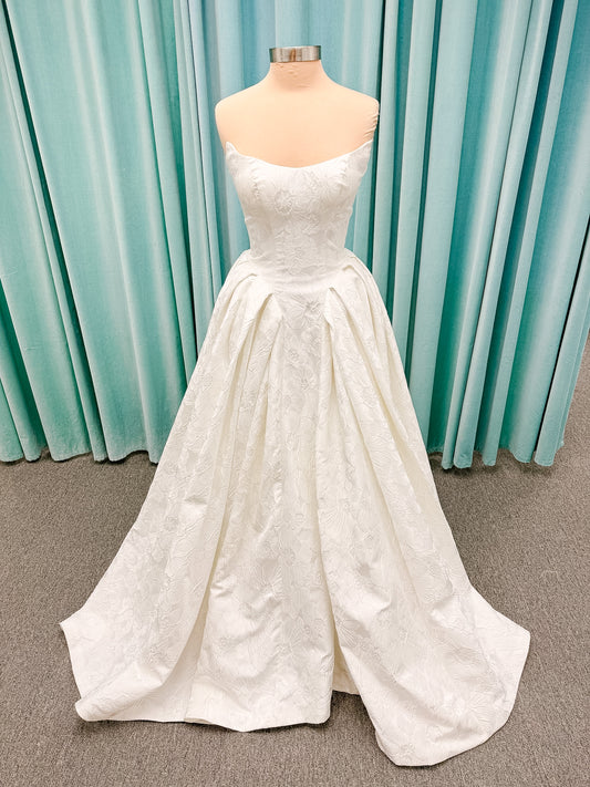 Haley Mai Bridal Lorea ballgown wedding dress on mannequin with strapless scoop neckline, seamed basque waist bodice, pleated ivory floral jacquard skirt, and long train