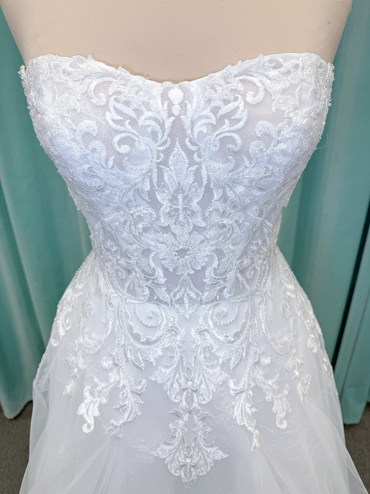 Moonlight Bridal J6972 Lunetta A-line wedding dress on mannequin with strapless soft sweetheart neckline, beaded lace appliques over tulle and Chantilly lace, drop waist, and scalloped applique sweep train