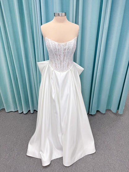 Moonlight Bridal J6998 Lusine A-line wedding dress on mannequin with strapless illusion scoop neckline, corded lace bodice with exposed boning, pearl-trimmed basque waist, full satin skirt with pockets, half lace-up back, and chapel train