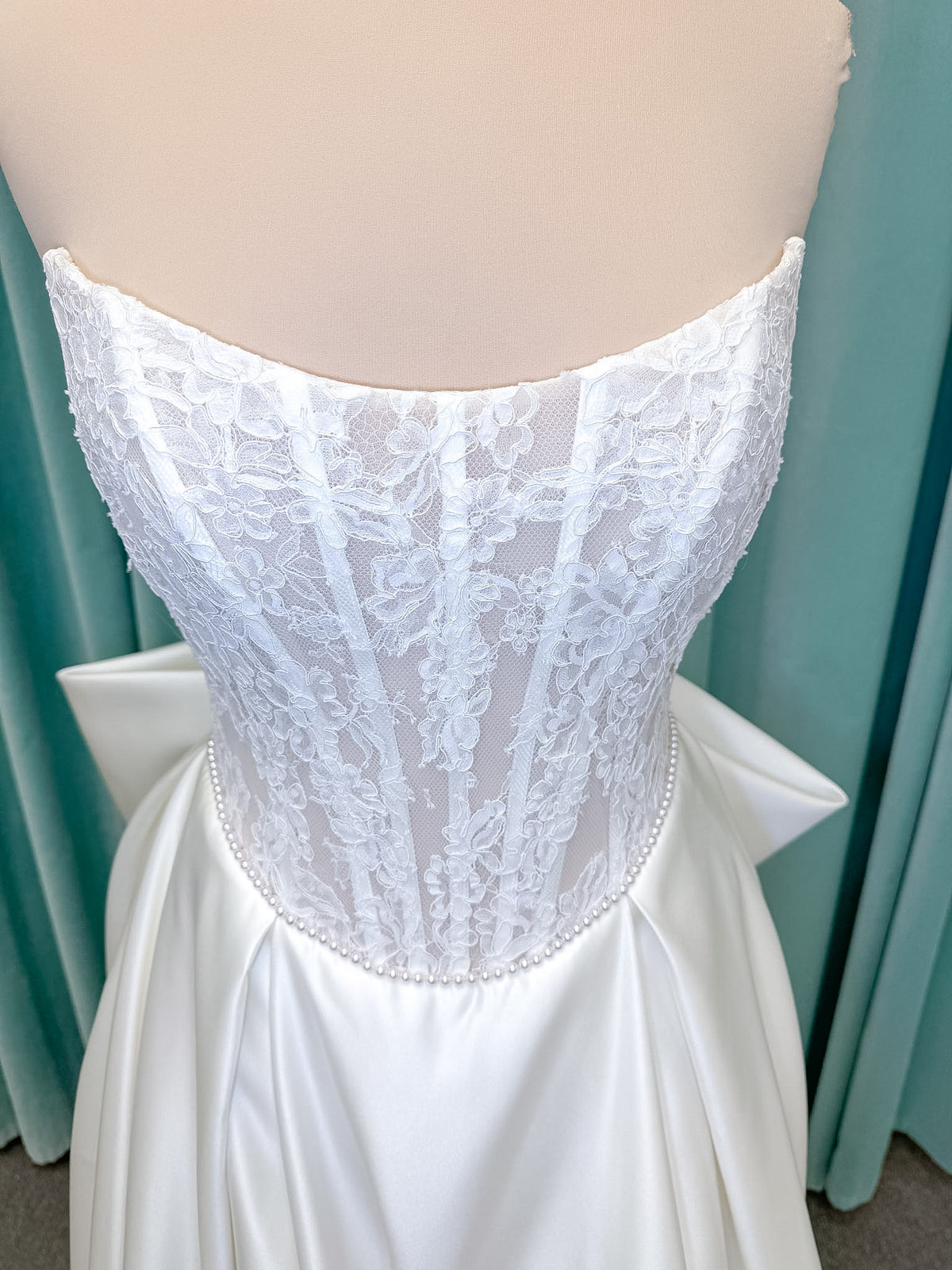 Moonlight Bridal J6998 Lusine A-line wedding dress on mannequin with strapless illusion scoop neckline, corded lace bodice with exposed boning, pearl-trimmed basque waist, full satin skirt with pockets, half lace-up back, and chapel train