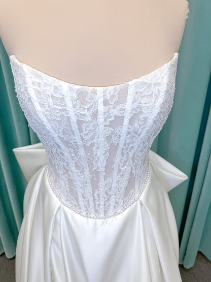 Moonlight Bridal J6998 Lusine A-line wedding dress on mannequin with strapless illusion scoop neckline, corded lace bodice with exposed boning, pearl-trimmed basque waist, full satin skirt with pockets, half lace-up back, and chapel train