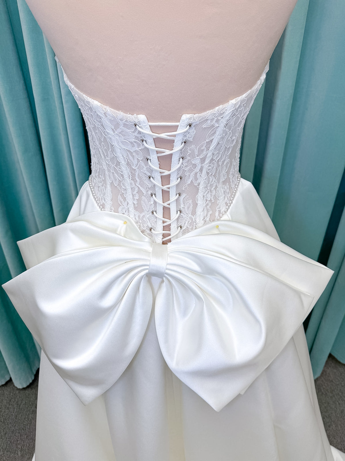 Moonlight Bridal J6998 Lusine A-line wedding dress on mannequin with strapless illusion scoop neckline, corded lace bodice with exposed boning, pearl-trimmed basque waist, full satin skirt with pockets, half lace-up back, and chapel train