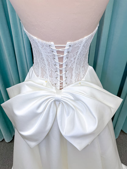 Moonlight Bridal J6998 Lusine A-line wedding dress on mannequin with strapless illusion scoop neckline, corded lace bodice with exposed boning, pearl-trimmed basque waist, full satin skirt with pockets, half lace-up back, and chapel train
