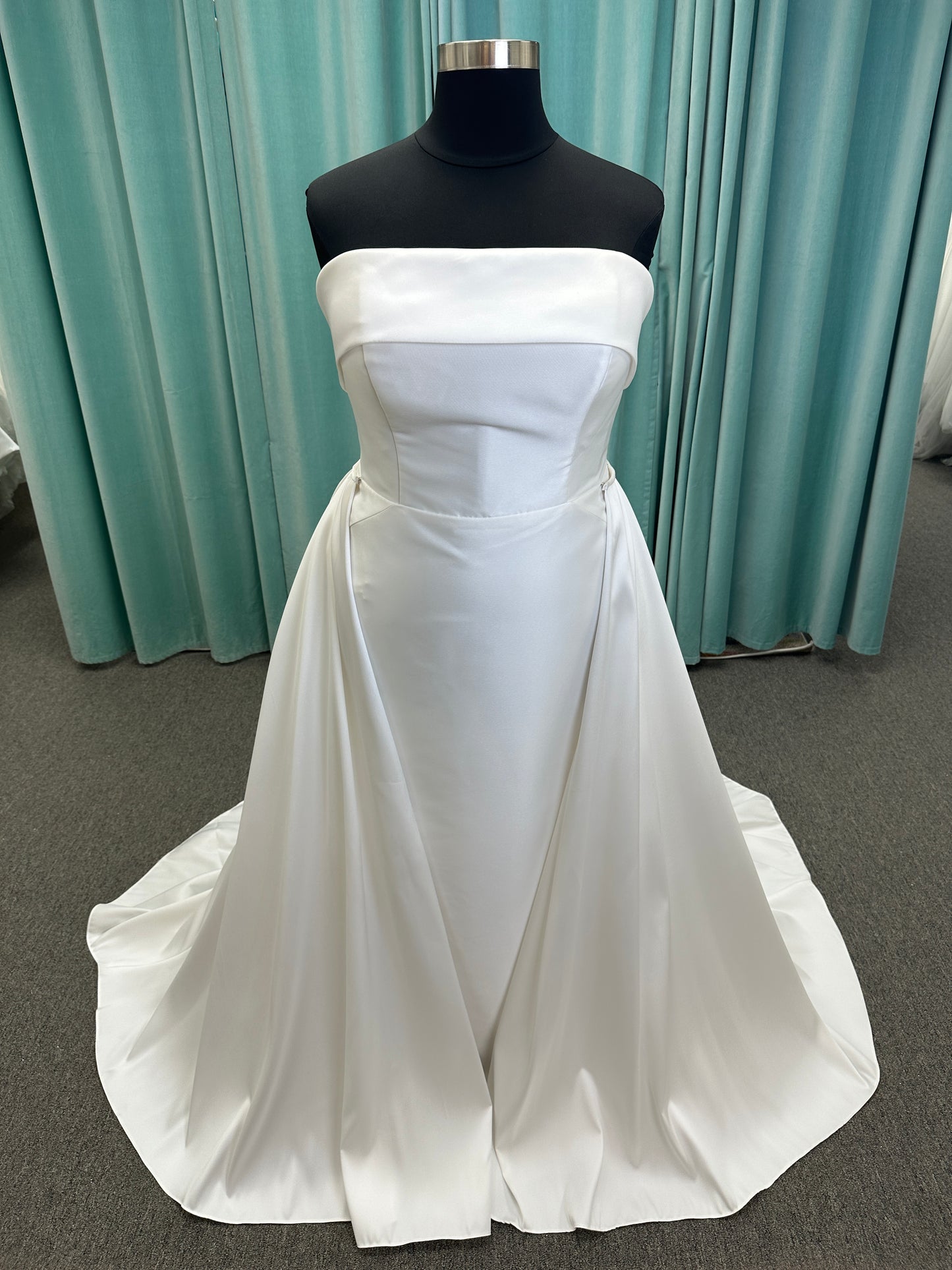 Oh Darling ivory strapless satin sheath wedding dress on mannequin with bandeau neckline, oversized statement bow at the waist, and voluminous overskirt.
