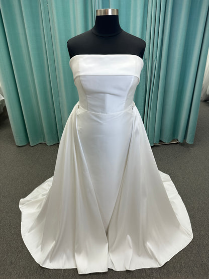 Oh Darling ivory strapless satin sheath wedding dress on mannequin with bandeau neckline, oversized statement bow at the waist, and voluminous overskirt.
