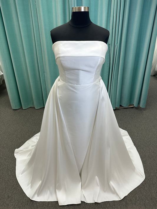 Oh Darling ivory strapless satin sheath wedding dress on mannequin with bandeau neckline, oversized statement bow at the waist, and voluminous overskirt.
