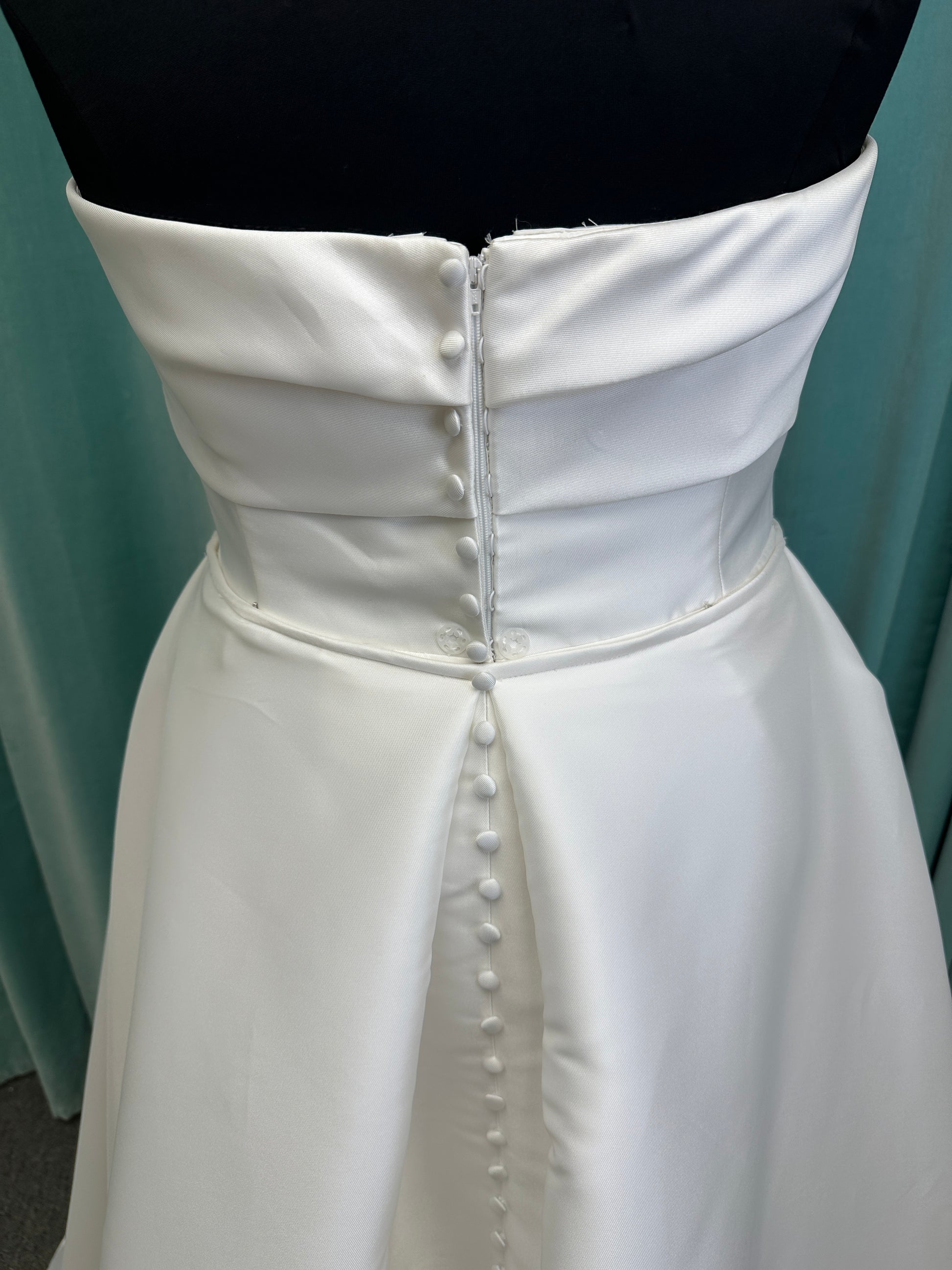 Oh Darling ivory strapless satin sheath wedding dress on mannequin with bandeau neckline, oversized statement bow at the waist, and voluminous overskirt.
