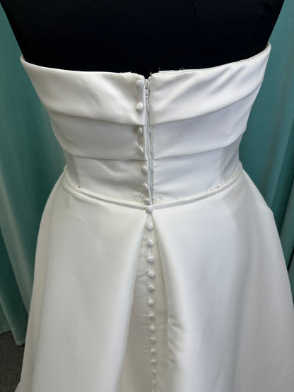 Oh Darling ivory strapless satin sheath wedding dress on mannequin with bandeau neckline, oversized statement bow at the waist, and voluminous overskirt.
