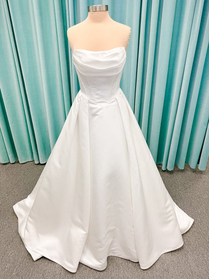 Stella York SY8084 ballgown wedding dress on mannequin with strapless ruched neckline, basque waist bodice, pleated satin skirt, and row of fabric-covered buttons