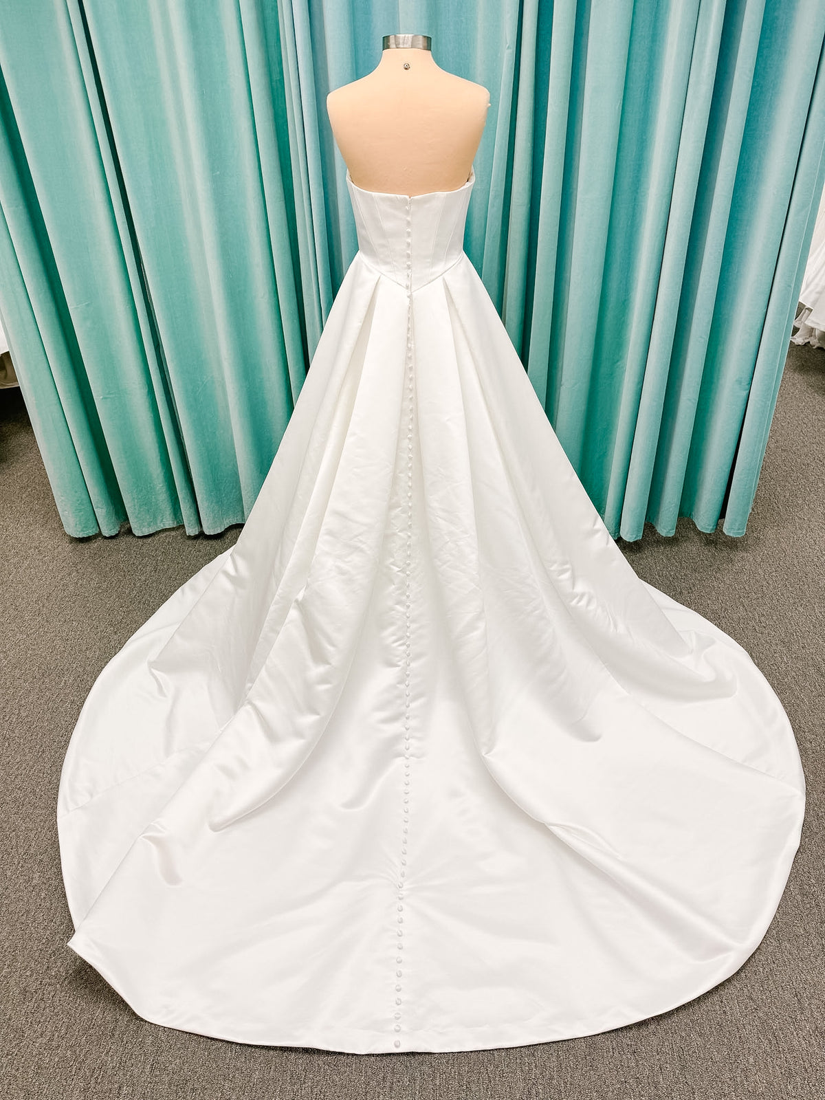 Stella York SY8084 ballgown wedding dress on mannequin with strapless ruched neckline, basque waist bodice, pleated satin skirt, and row of fabric-covered buttons