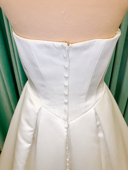 Stella York SY8084 ballgown wedding dress on mannequin with strapless ruched neckline, basque waist bodice, pleated satin skirt, and row of fabric-covered buttons