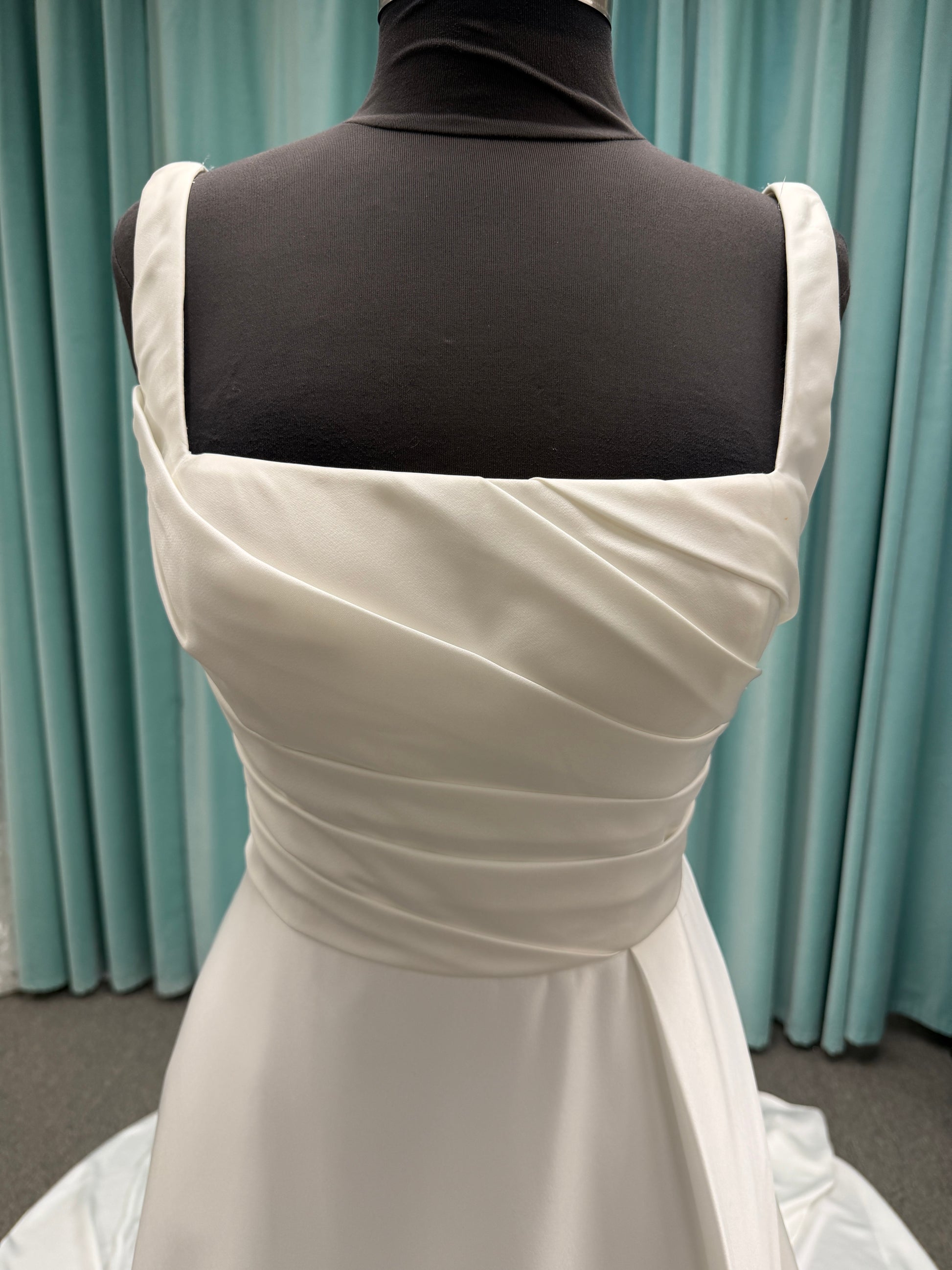 Stella York SY8110 ivory satin A-line wedding dress on mannequin with square neckline, shoulder straps, ruched bodice, and scoop back with fabric-covered buttons.
