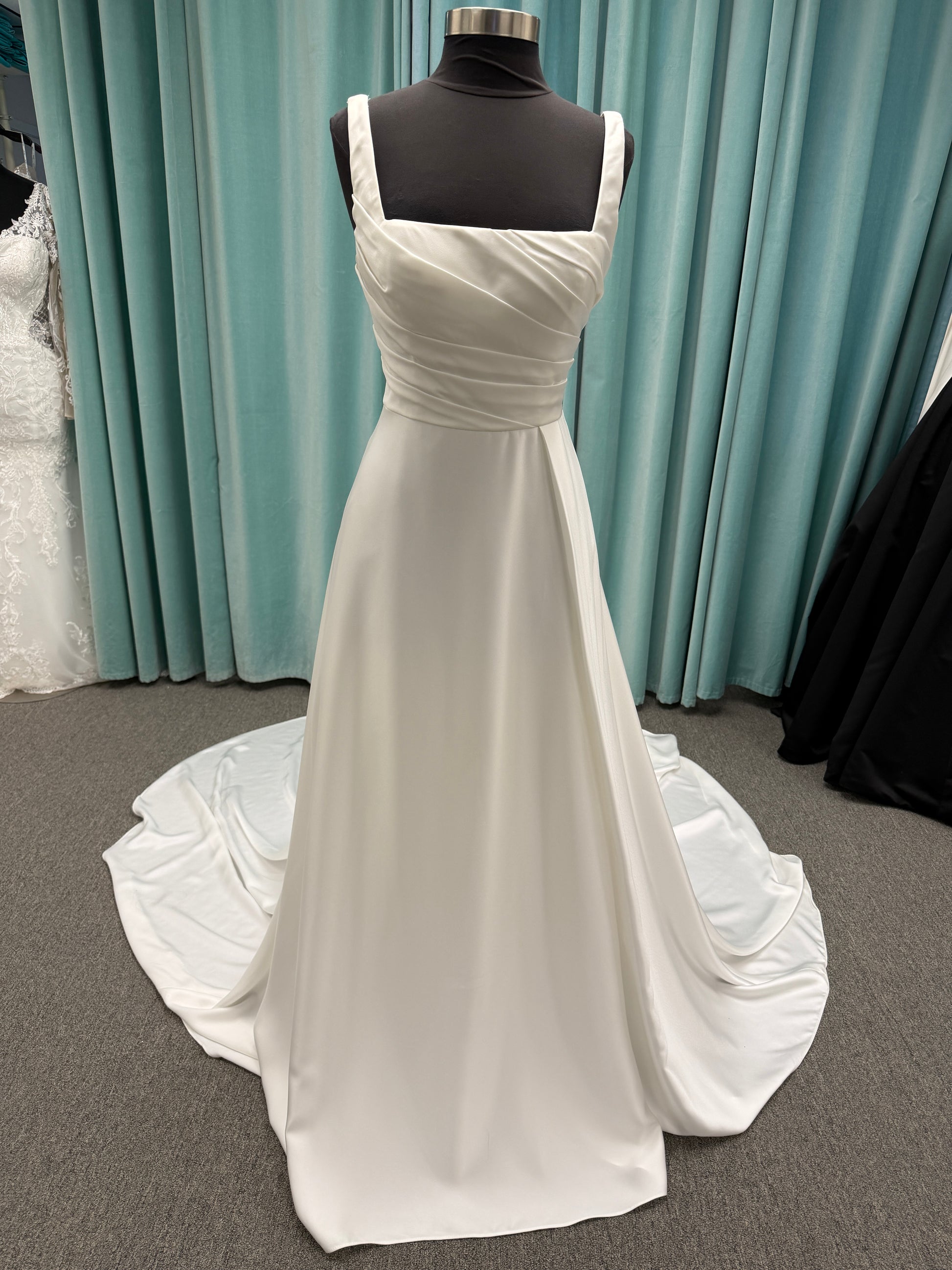 Stella York SY8110 ivory satin A-line wedding dress on mannequin with square neckline, shoulder straps, ruched bodice, and scoop back with fabric-covered buttons.
