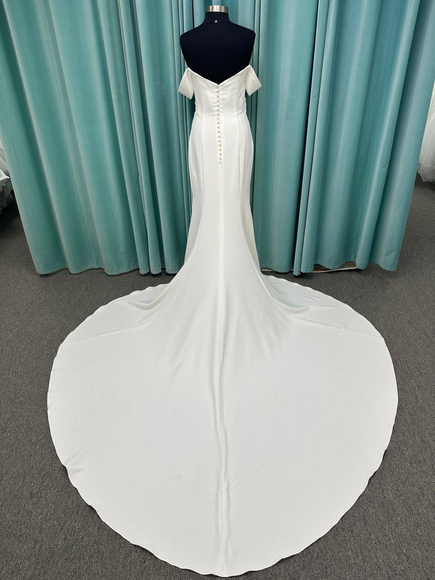 Tai ivory off-the-shoulder crepe sheath wedding dress on mannequin with draped bodice, front slit, and cathedral train.
