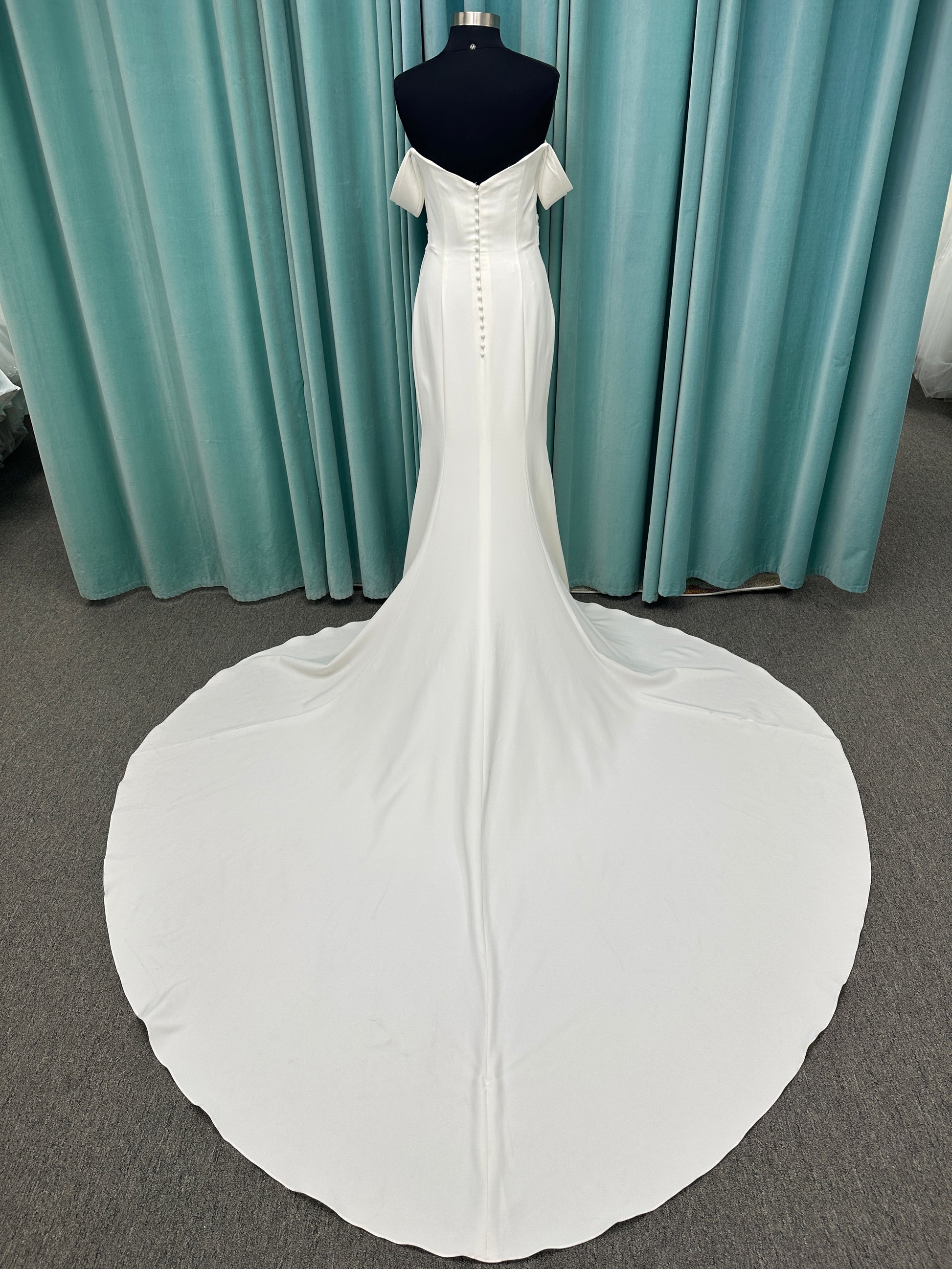 Tai ivory off-the-shoulder crepe sheath wedding dress on mannequin with draped bodice, front slit, and cathedral train.
