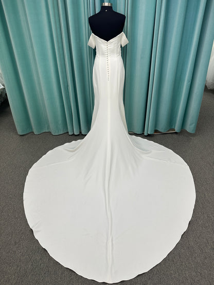 Tai ivory off-the-shoulder crepe sheath wedding dress on mannequin with draped bodice, front slit, and cathedral train.
