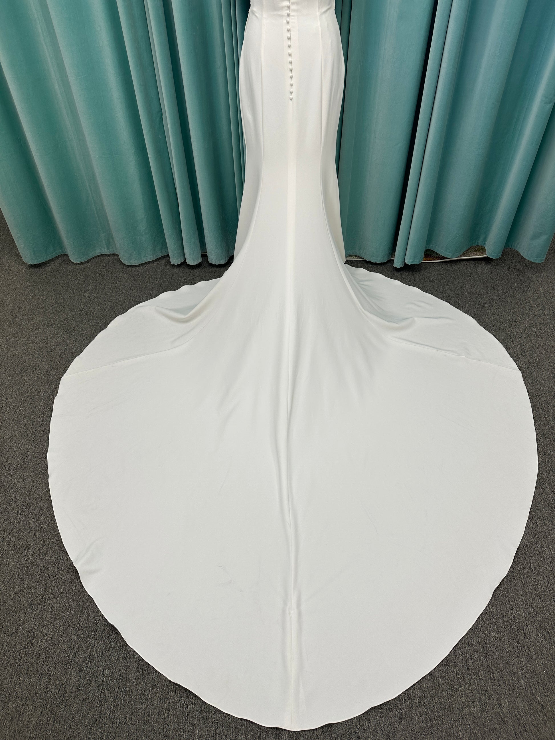 Tai ivory off-the-shoulder crepe sheath wedding dress on mannequin with draped bodice, front slit, and cathedral train.
