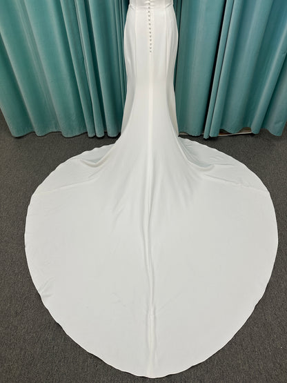 Tai ivory off-the-shoulder crepe sheath wedding dress on mannequin with draped bodice, front slit, and cathedral train.
