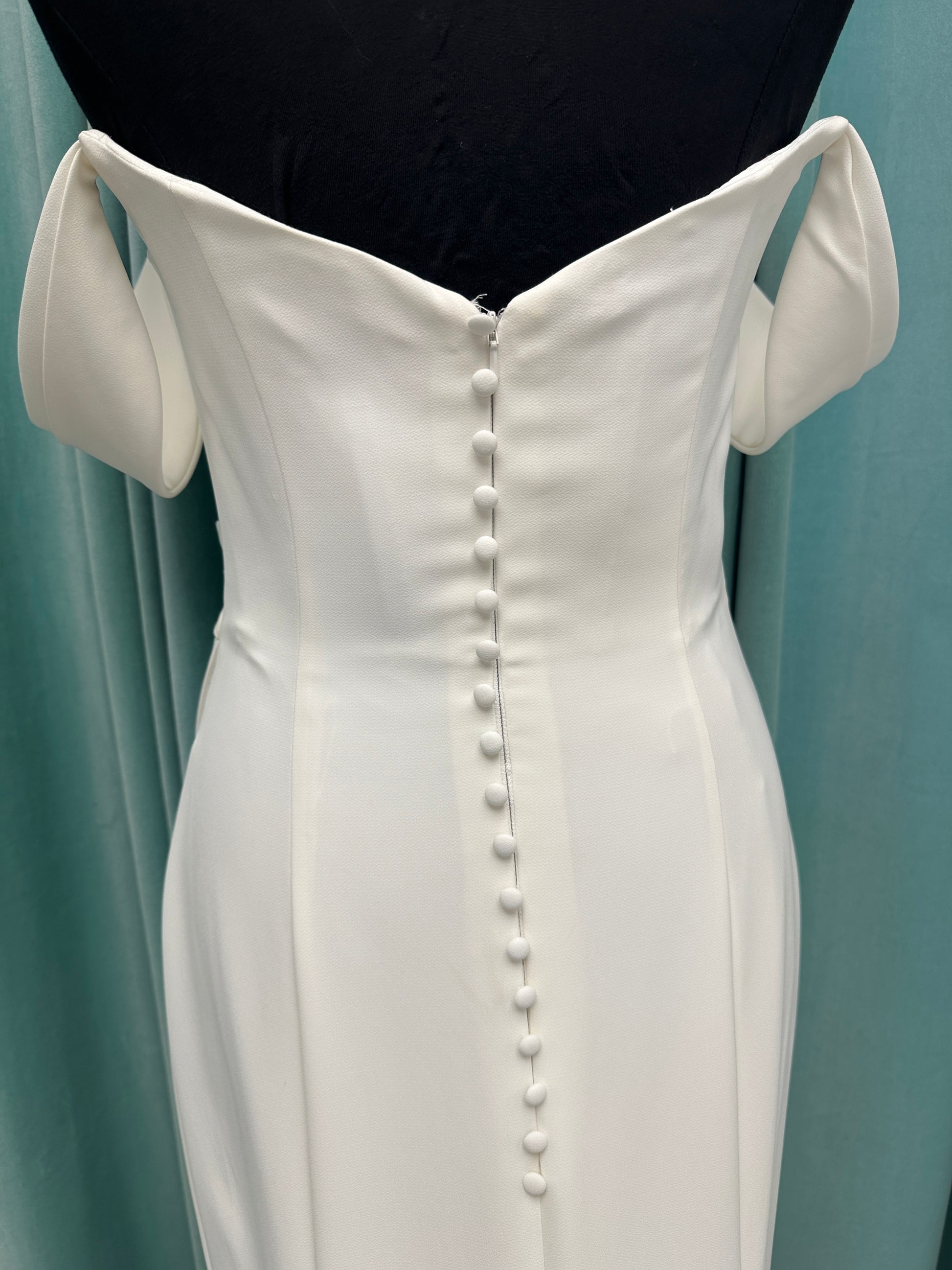 Tai ivory off-the-shoulder crepe sheath wedding dress on mannequin with draped bodice, front slit, and cathedral train.
