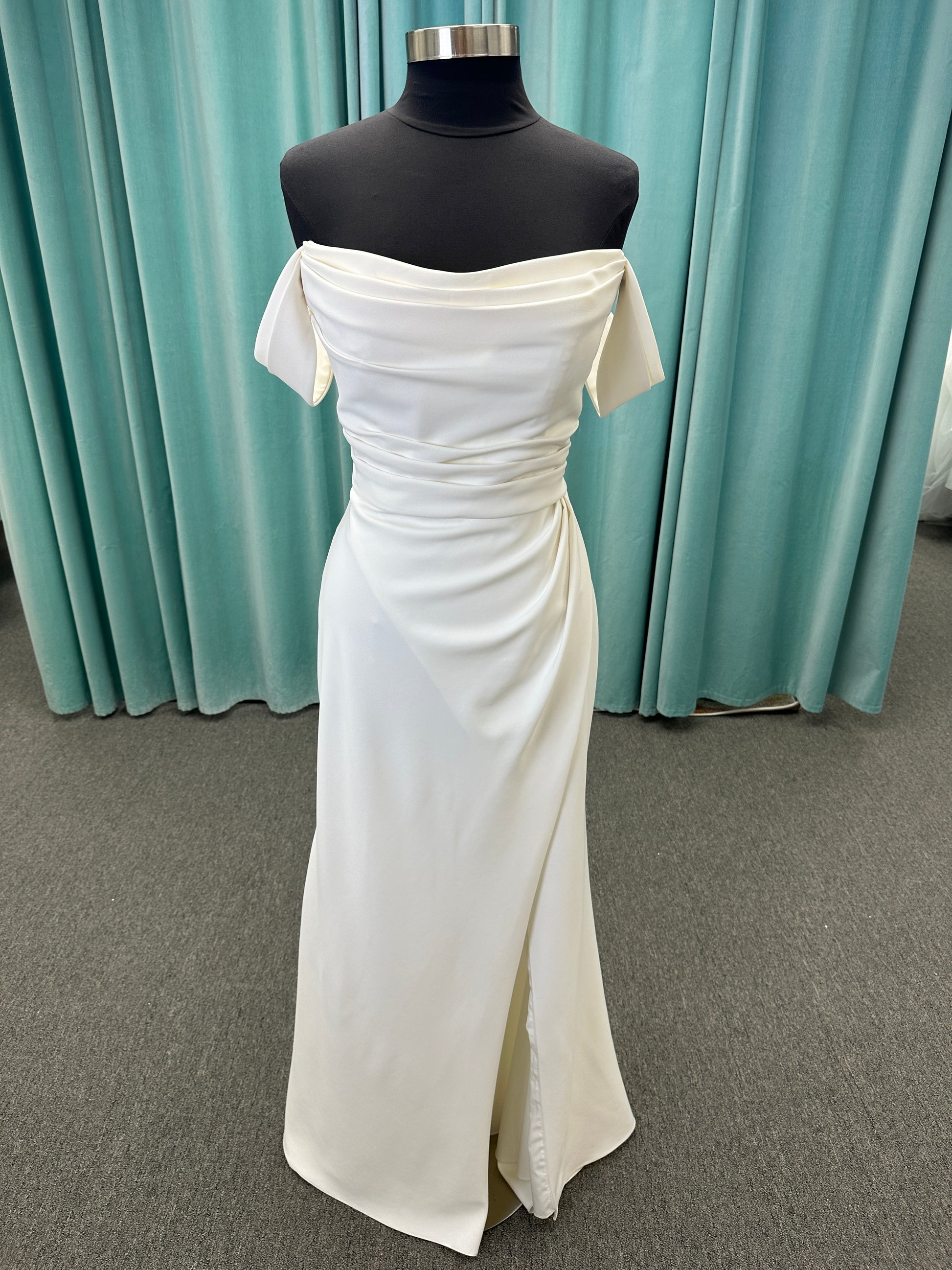 Tai ivory off-the-shoulder crepe sheath wedding dress on mannequin with draped bodice, front slit, and cathedral train.
