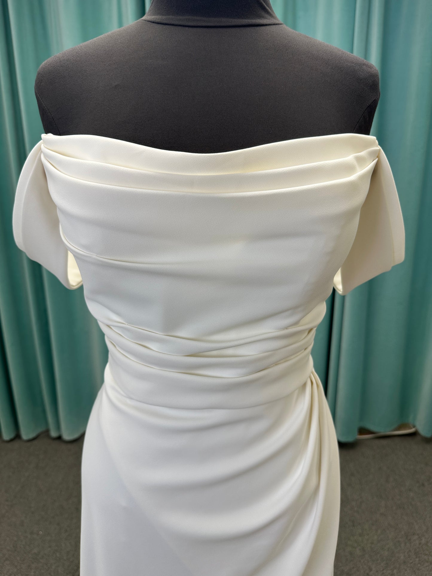 Tai ivory off-the-shoulder crepe sheath wedding dress on mannequin with draped bodice, front slit, and cathedral train.
