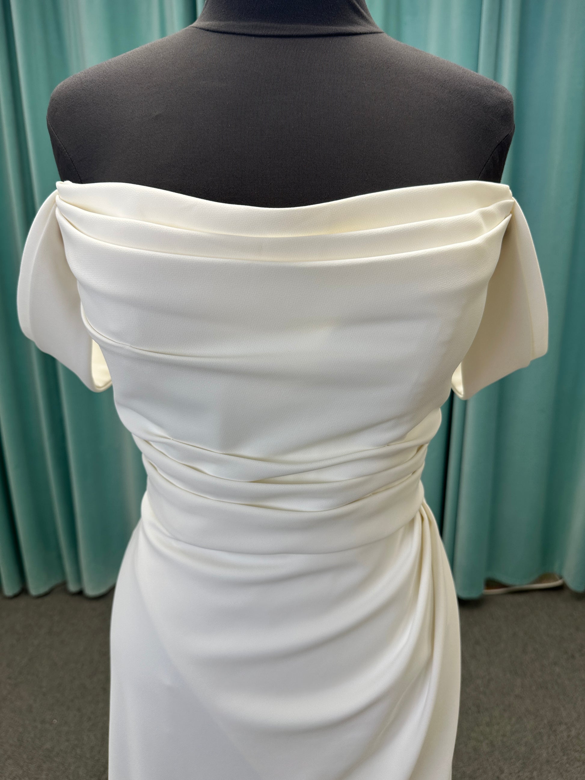 Tai ivory off-the-shoulder crepe sheath wedding dress on mannequin with draped bodice, front slit, and cathedral train.
