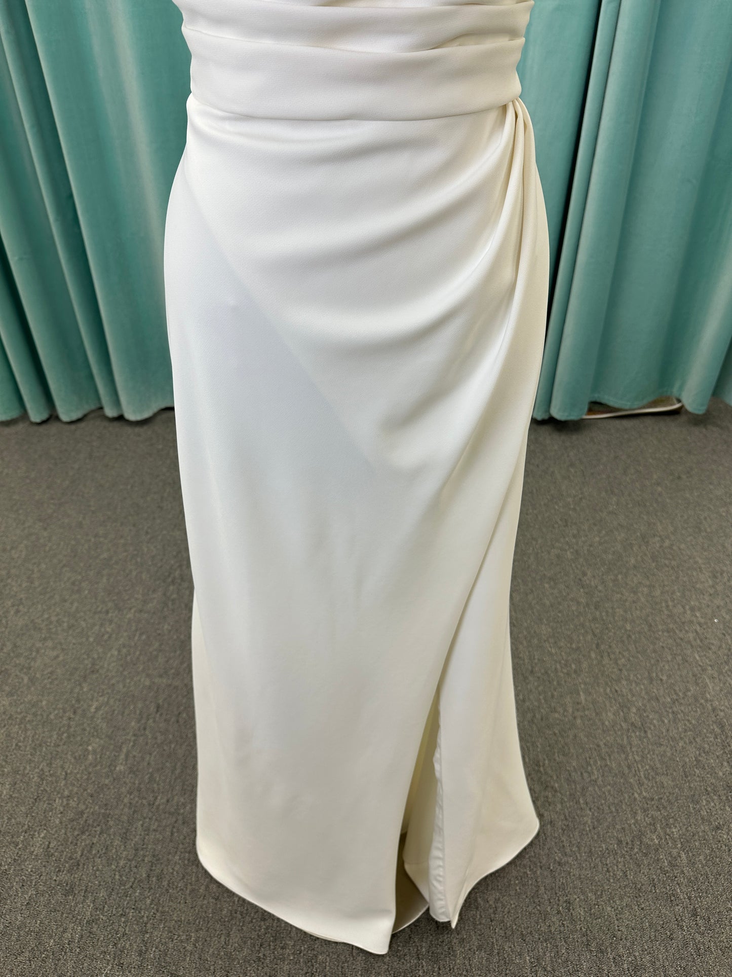 Tai ivory off-the-shoulder crepe sheath wedding dress on mannequin with draped bodice, front slit, and cathedral train.
