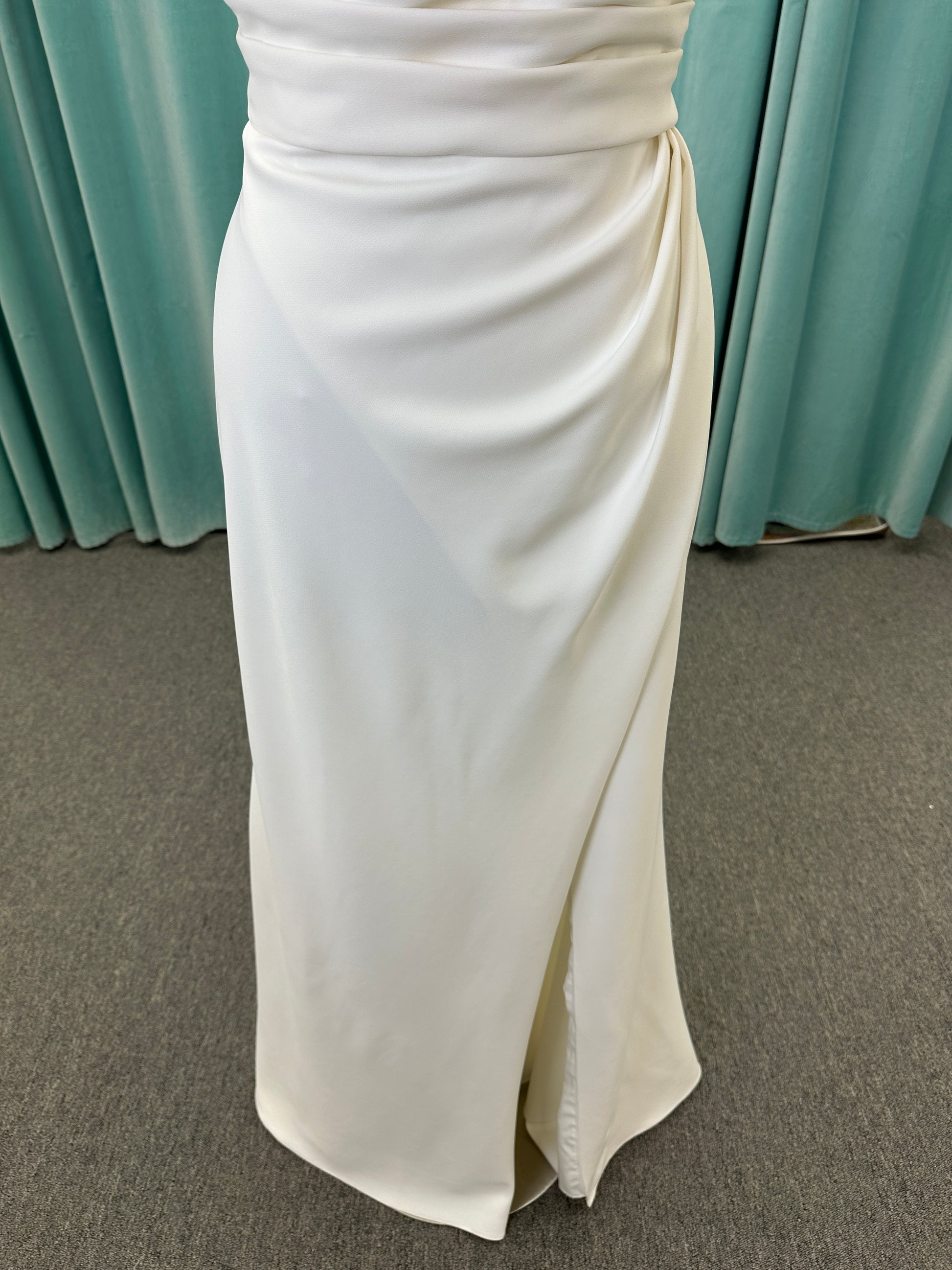 Tai ivory off-the-shoulder crepe sheath wedding dress on mannequin with draped bodice, front slit, and cathedral train.
