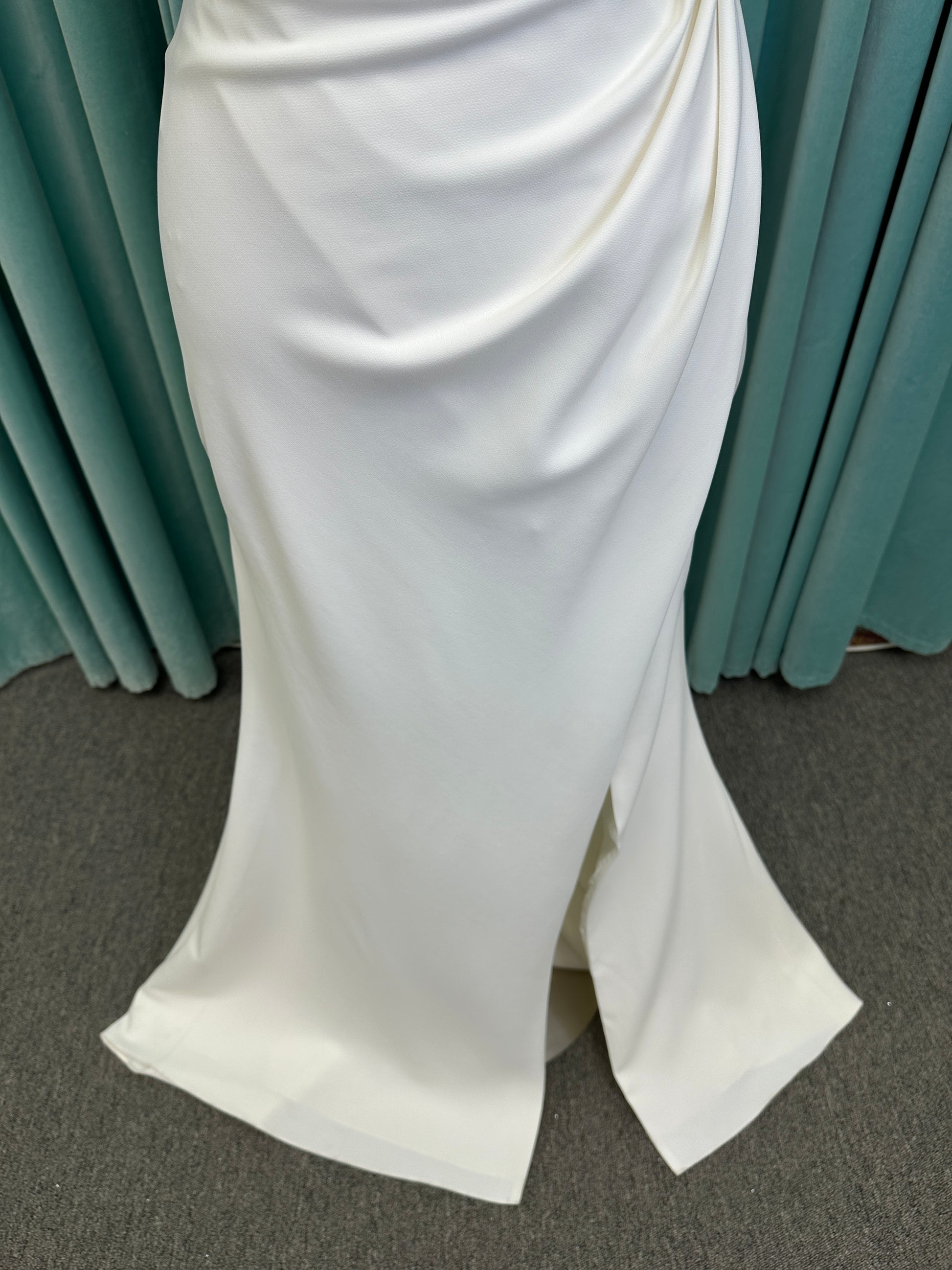 Tai ivory off-the-shoulder crepe sheath wedding dress on mannequin with draped bodice, front slit, and cathedral train.
