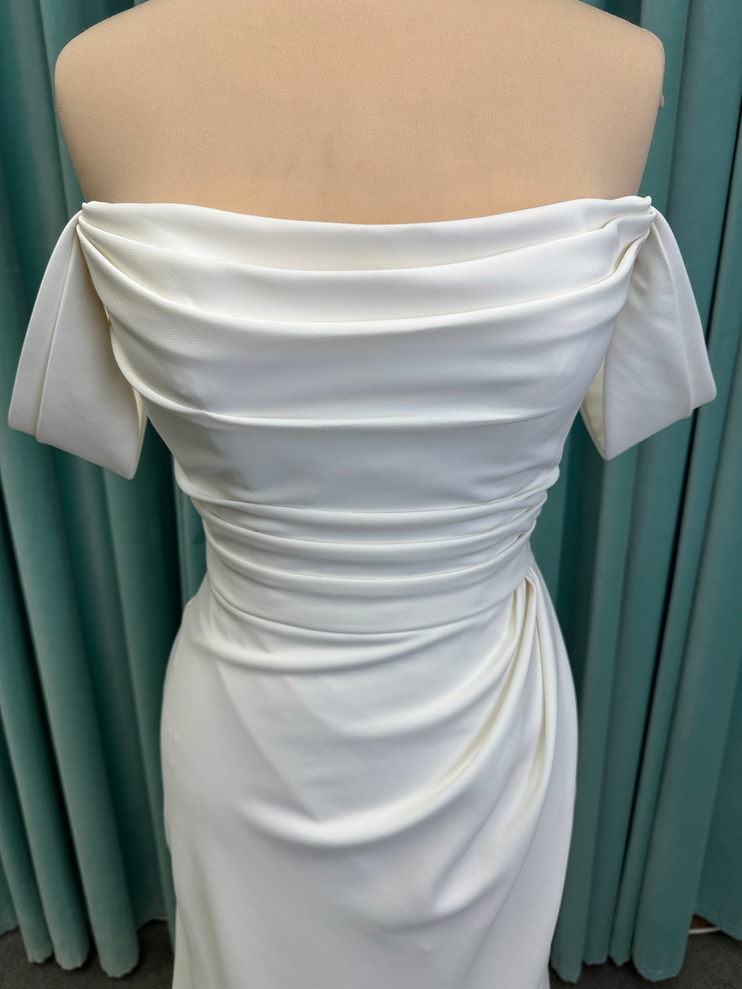 Tai ivory off-the-shoulder crepe sheath wedding dress on mannequin with draped bodice, front slit, and cathedral train.
