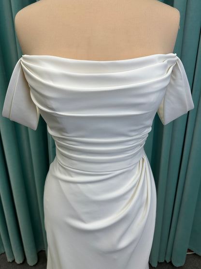 Tai ivory off-the-shoulder crepe sheath wedding dress on mannequin with draped bodice, front slit, and cathedral train.
