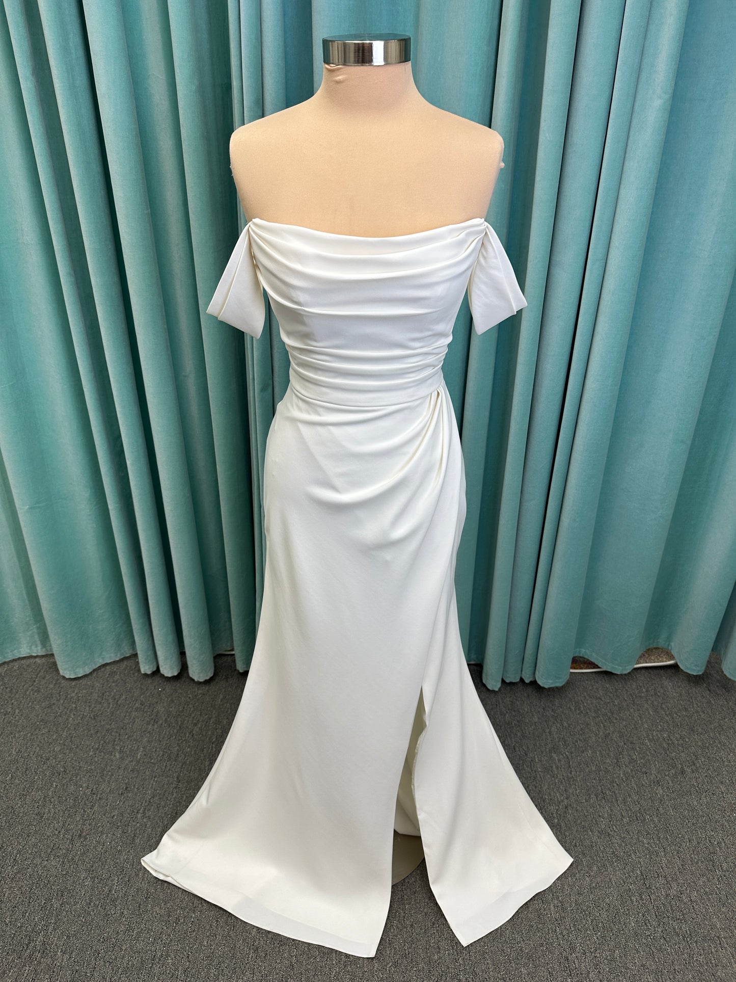 Tai ivory off-the-shoulder crepe sheath wedding dress on mannequin with draped bodice, front slit, and cathedral train.
