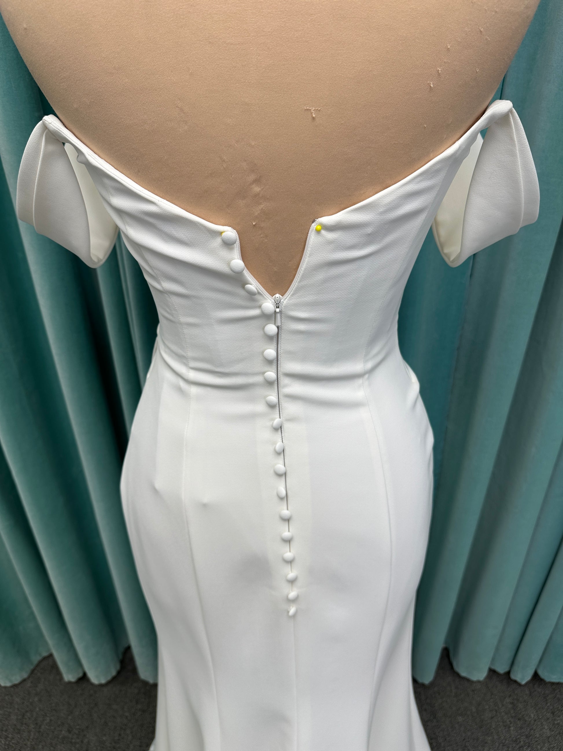 Tai ivory off-the-shoulder crepe sheath wedding dress on mannequin with draped bodice, front slit, and cathedral train.
