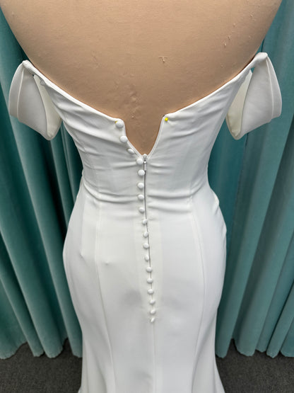 Tai ivory off-the-shoulder crepe sheath wedding dress on mannequin with draped bodice, front slit, and cathedral train.
