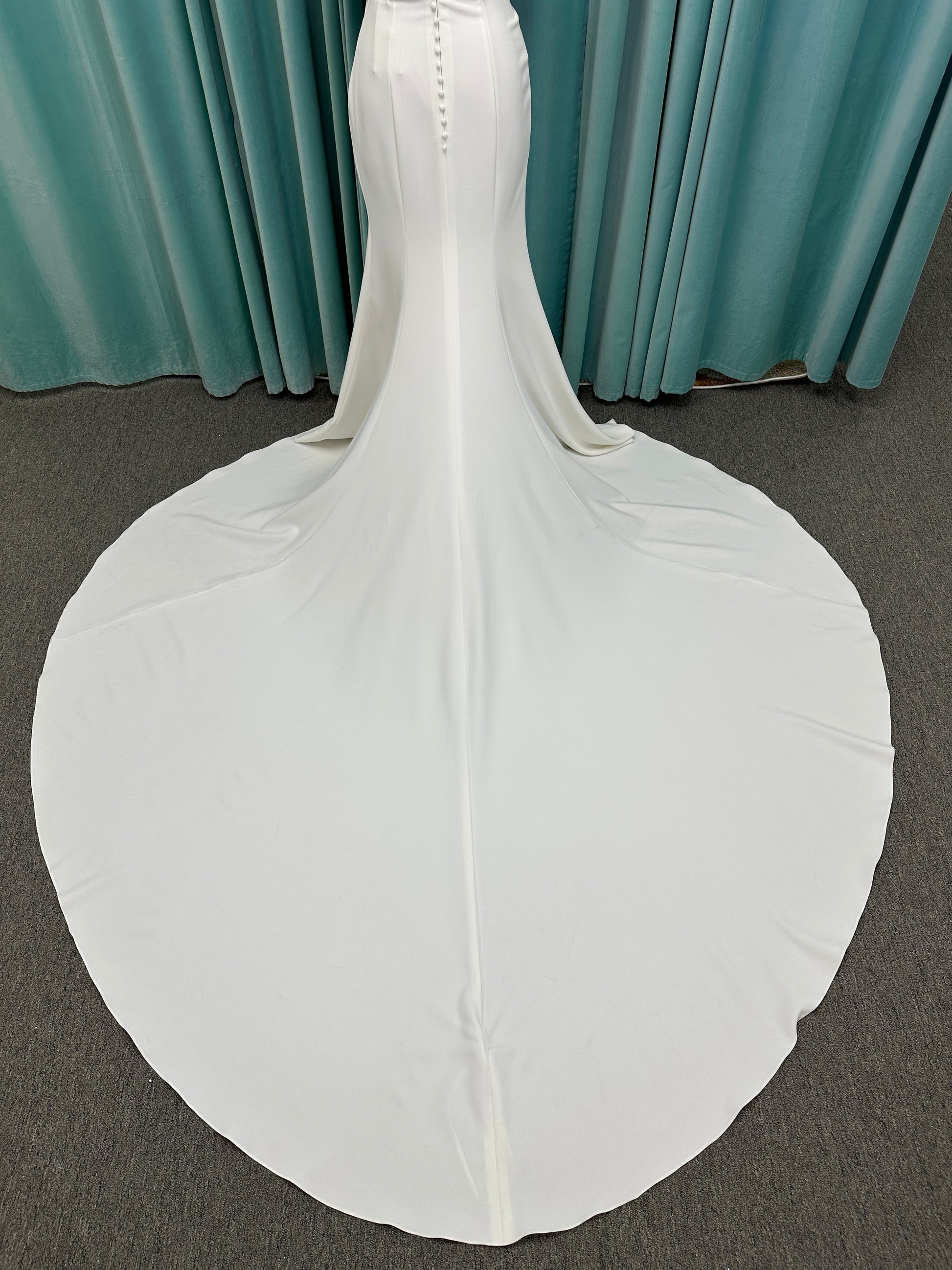 Tai ivory off-the-shoulder crepe sheath wedding dress on mannequin with draped bodice, front slit, and cathedral train.
