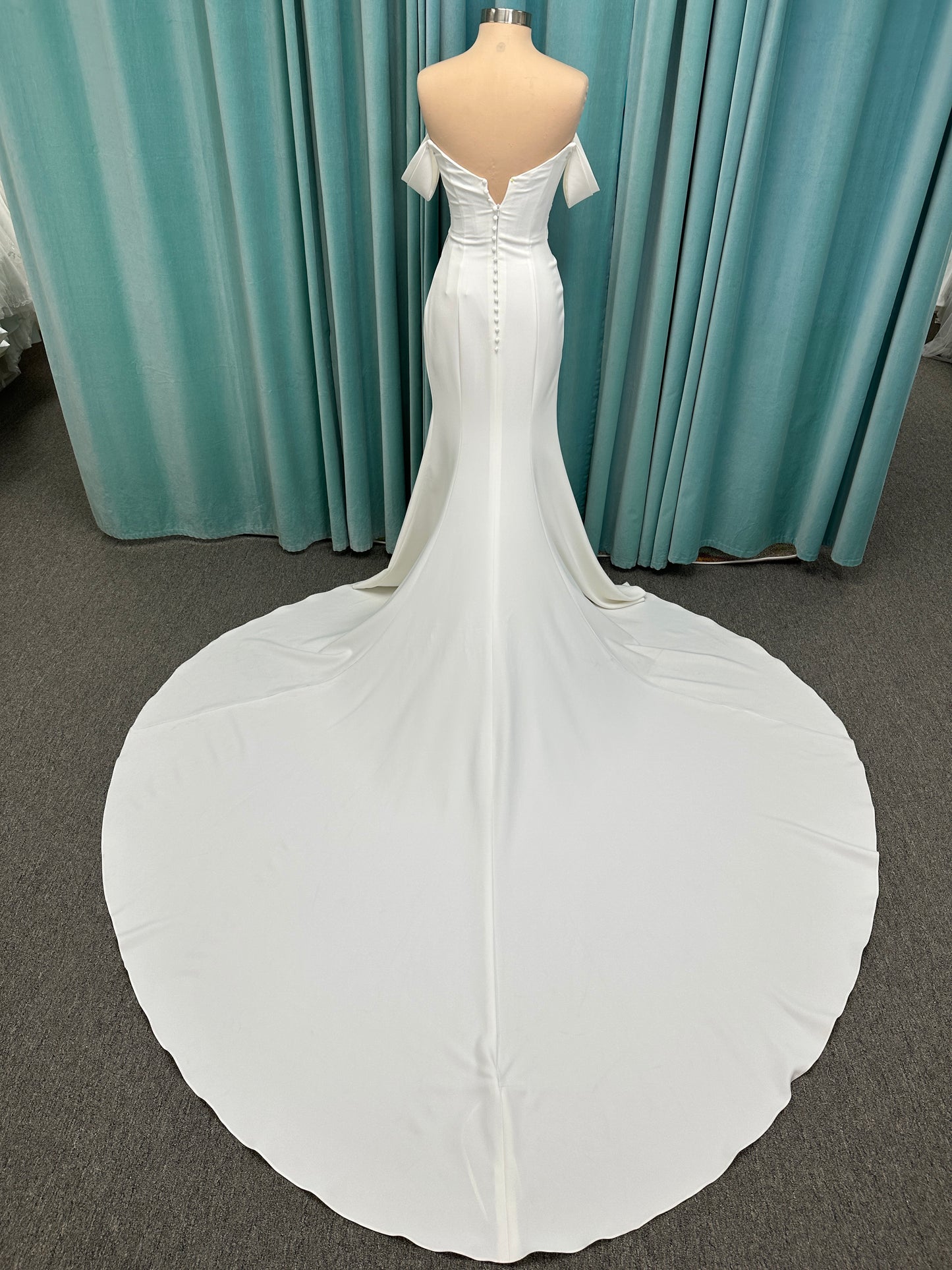 Tai ivory off-the-shoulder crepe sheath wedding dress on mannequin with draped bodice, front slit, and cathedral train.
