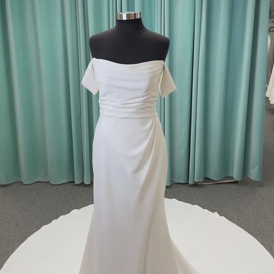 Tai ivory off-the-shoulder crepe sheath wedding dress on mannequin with draped bodice, front slit, and cathedral train.
